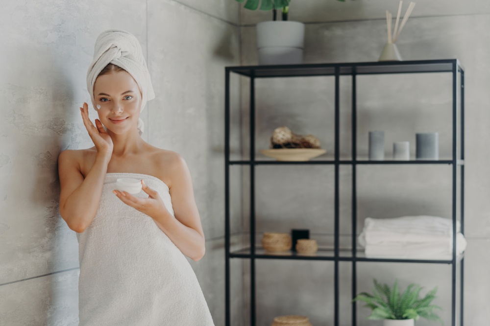 Horizontal shot of young healthy female model uses cream moisturizer, holds jar of cosmetic product, wrapped in towel after showering, stands near wall in cozy bathroom. Skin care procedures