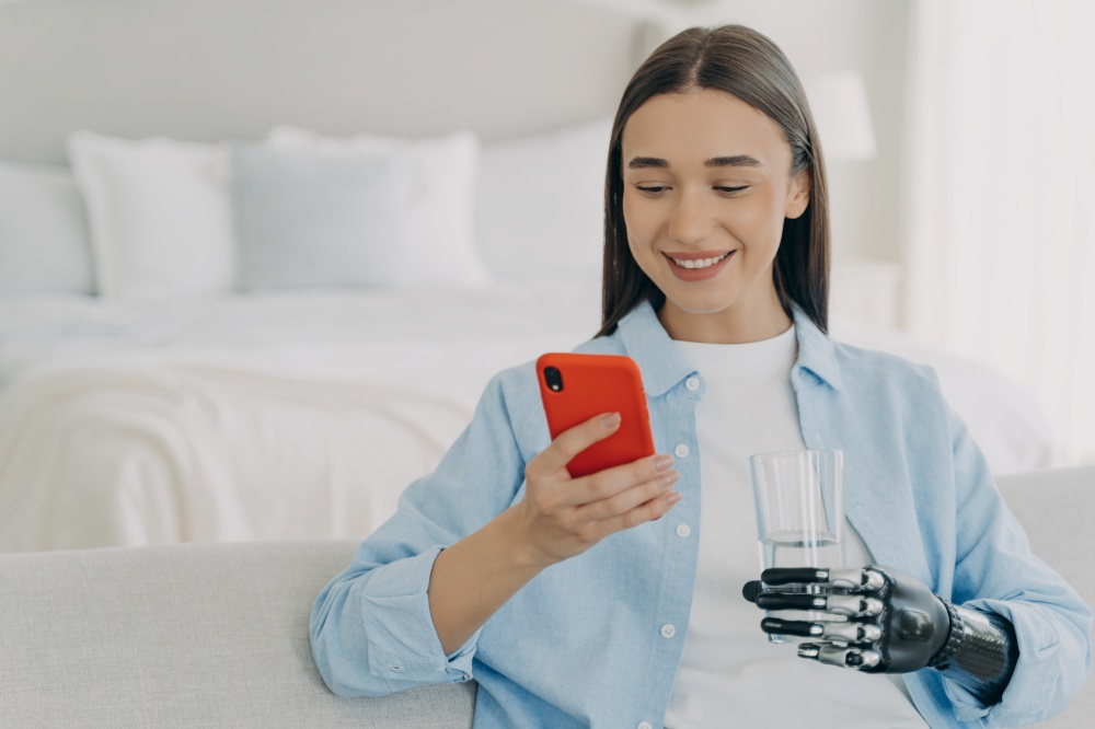 Smiling young disabled woman holding smartphone and glass of pure water. Female with bionic prosthetic arm using mobile phone apps, chatting in social networks, shopping online at home.. Disabled girl with bionic prosthetic arm using mobile phone apps at home, holding glass of water