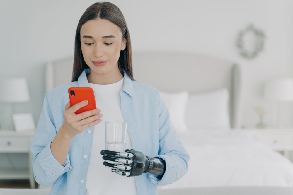 Young disabled girl holds smartphone and glass of water uses bionic prosthesis, chatting in social network, shopping online. Female with robotic prosthetic hand enjoy using mobile phone apps.. Young disabled girl holds smartphone and glass of water in hands using high tech bionic prosthesis