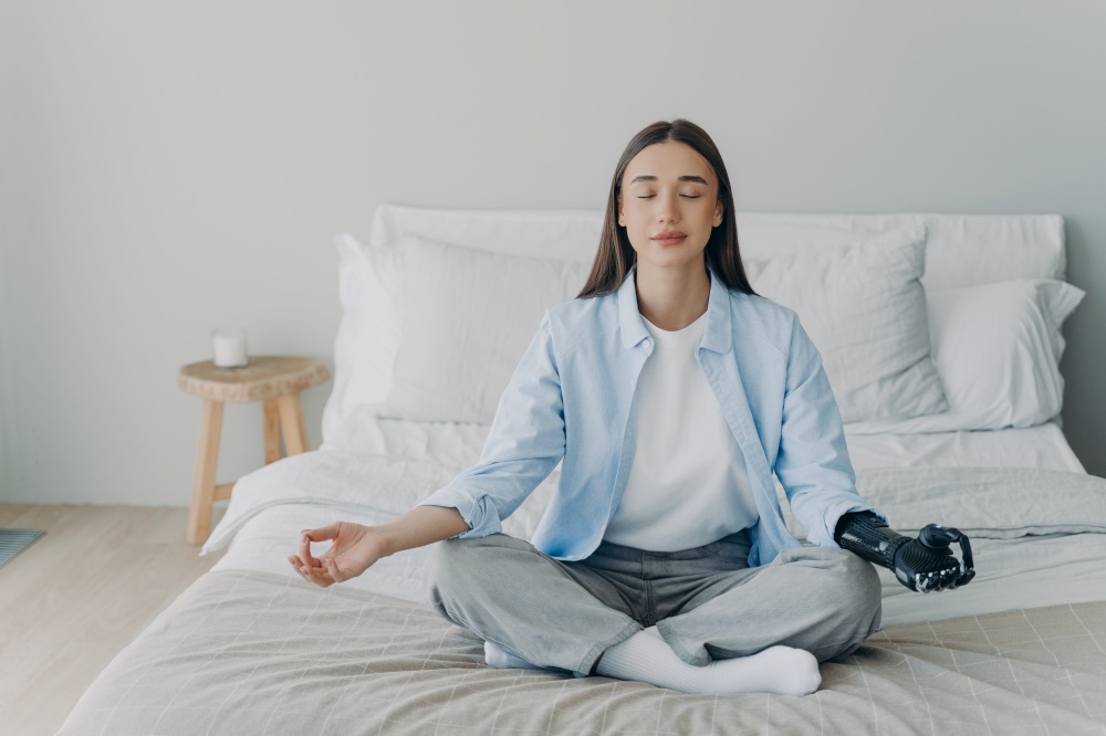 Calm disabled young caucasian girl with bionic arm prosthesis relaxing in lotus pose sitting on bed. Female with disability practicing yoga, meditating, breathing, reducing stress.. Disabled girl with bionic arm prosthesis practice yoga, meditating in lotus pose on bed at home