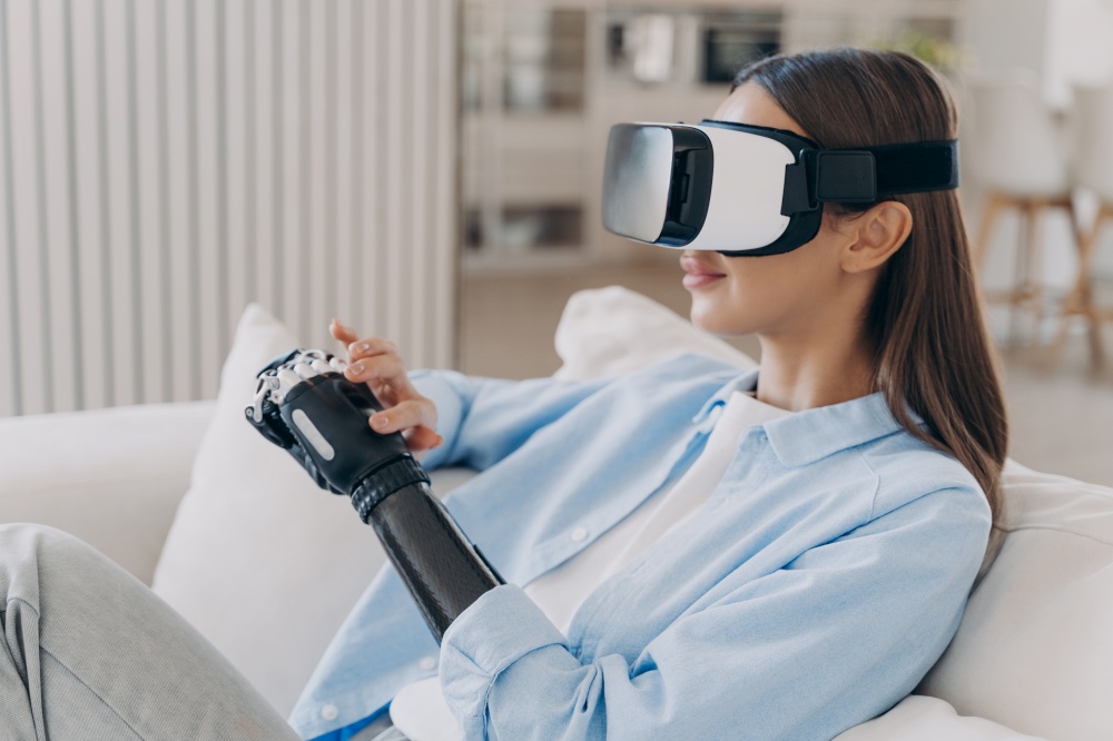Modern disabled girl in VR glasses interacts with augmented reality, touching her high tech bionic prosthetic arm, sitting on sofa at home. Disability and medical future technologies concept.. Disabled girl in VR glasses interacts with augmented reality, touches her high tech prosthetic arm
