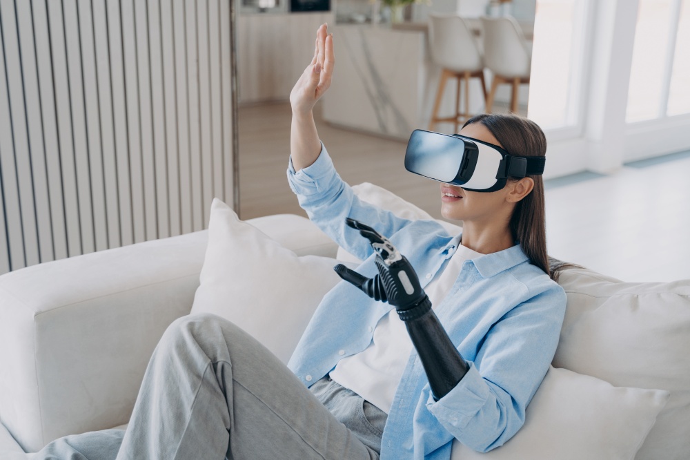 Modern young disabled woman wearing virtual reality glasses touching 3d objects in cyberspace, sitting on sofa. Girl with disability in vr goggles learning to use artificial limb, playing video game.. Modern disabled girl in virtual reality glasses playing video game, learning to use prosthetic arm