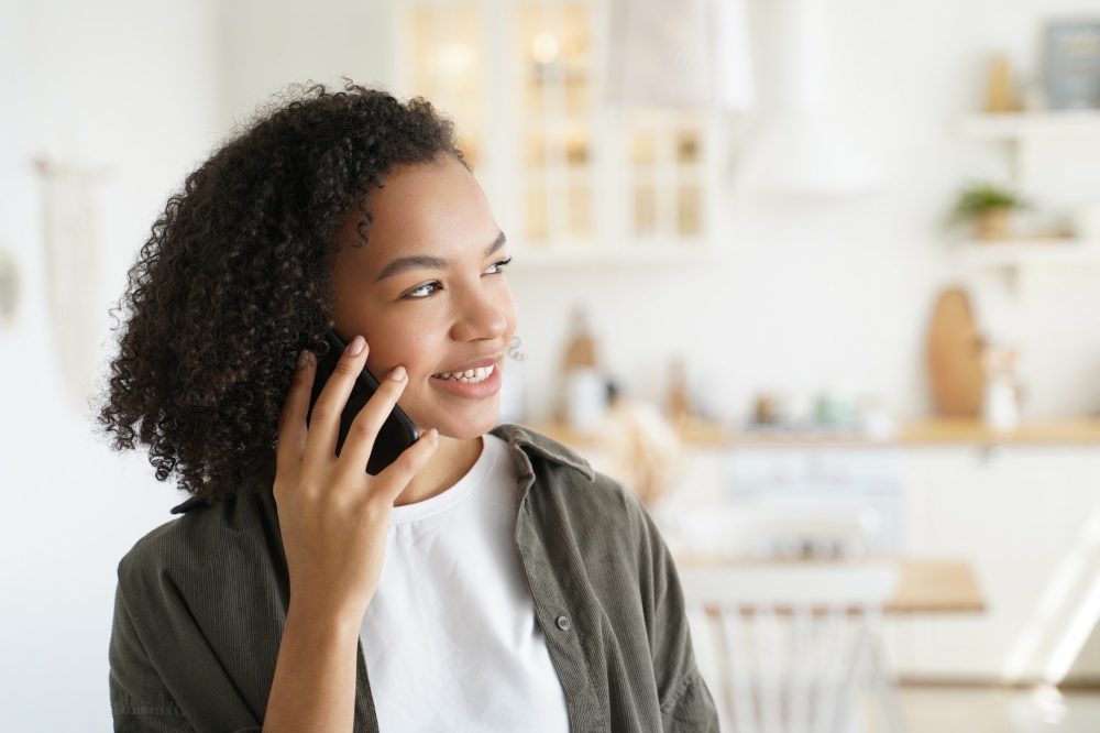 Friendly african american teen girl talking on smartphone, makes phone call from home. Smiling young lady with afro hairs enjoying pleasant mobile conversation with friends indoors.. Friendly african american teen girl talking on smartphone, makes mobile phone call from home