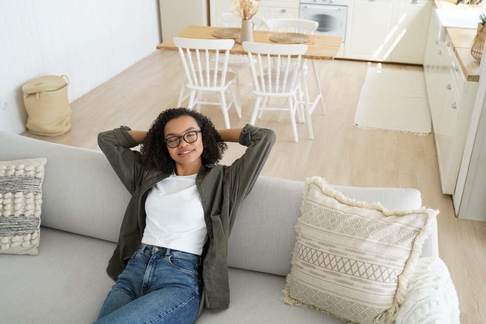 Relaxed biracial young girl in glasses rest, dreaming, sitting on couch in cozy living room at home. Calm woman relaxing on comfortable sofa with hands behind head, enjoying lazy break.. Relaxed biracial young girl in glasses rest, dreaming, sitting on couch in cozy living room at home