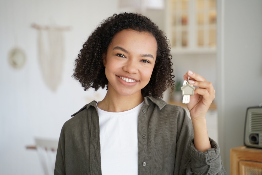Smiling mixed race teen girl tenant or homeowner holds keys to new home apartment, happy young biracial lady renter holding house key, standing indoors. Rental housing, mortgage advertisement.. Smiling mixed race teen girl tenant or homeowner holds keys to new home. Rental housing, mortgage