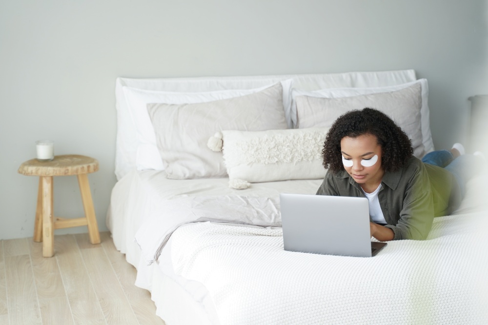 African american teen girl with under eye patches working on laptop lying on bed at home during daily skincare routine. Biracial young lady shopping online on computer while morning beauty procedure.. Biracial teen girl with under eye patches shopping online at laptop in bedroom while beauty routine