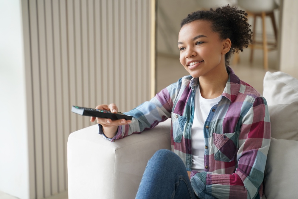 Smiling mixed race girl changes TV channel with remote control, watching television series on sofa at home. Modern young teen lady watches movie, relaxing on couch, enjoying leisure time.. Smiling mixed race girl changes TV channel with remote control, watching television series on sofa