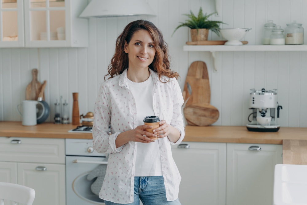 Smiling girl holding paper cup of coffee standing in modern cozy kitchen, looking at camera. Happy female housewife homeowner holds takeaway delivery hot drink to go at home. Domestic life. Smiling girl hold paper cup of coffee, standing in modern cozy kitchen look at camera. Domestic life