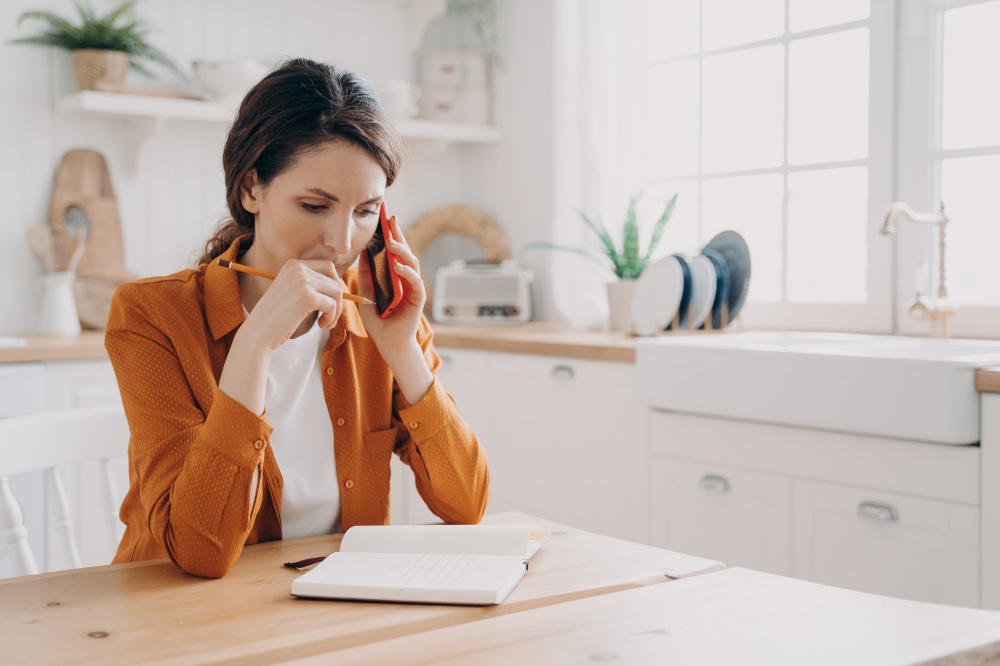 Puzzled woman making phone call, taking notes sitting at kitchen table. Pensive female freelance worker consulting client or discussing project with colleague at home. Remote job concept.. Female freelance worker making phone call, discussing work project in kitchen at home. Remote job