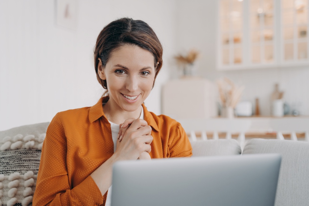 Smiling woman looks at laptop screen, reads email or message on social network with good news or online store offer at home. Happy girl housewife watching movie or series, touched with tender moment.. Touched woman looks at laptop screen, reads email, message on social network with good news at home