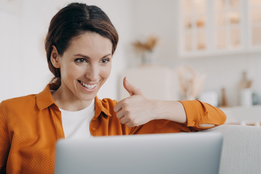 Smiling hispanic woman showing thumb up gesture looking at laptop screen, recommending online service. Satisfied female student gives recommendation to distant education, making video call at home.. Female shows thumb up gesture, looking at laptop screen, recommends online service, makes video call