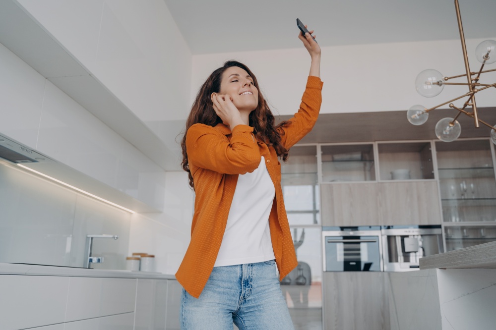Happy woman holding smartphone, dancing alone in kitchen at home. Smiling female with closed eyes dances listening to favorite music on phone, moving to song, enjoying musical sound.. Happy girl listening to music, holding phone dances to song alone in kitchen at home. Musical app ad