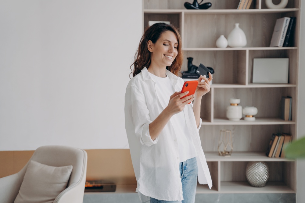 Smiling female holding bank credit card and smartphone in modern home interior. Woman paying for purchases in online store using banking service, makes secure cashless payment at home. E-commerce.. Happy woman holding bank credit card, smartphone, uses online banking services at home. E-commerce