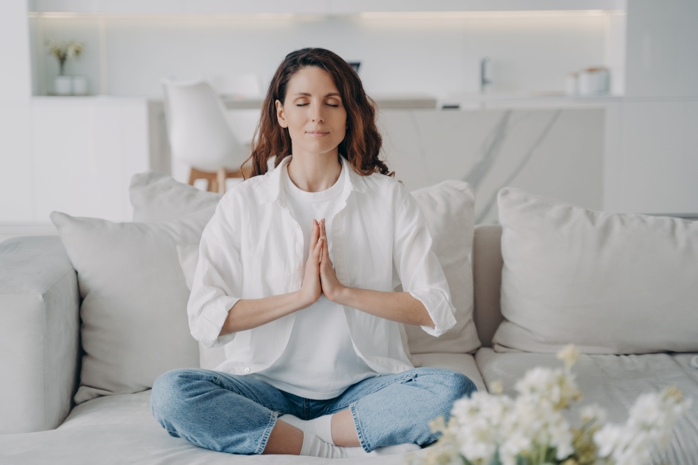 Peaceful spanish woman relaxing in lotus pose on comfortable couch at home. Calm female enjoying practicing yoga breathing exercises, meditation, sitting on sofa. Reducing stress, wellness concept.. Female practicing yoga breathing exercises on couch at home. Reducing stress, wellness, meditation