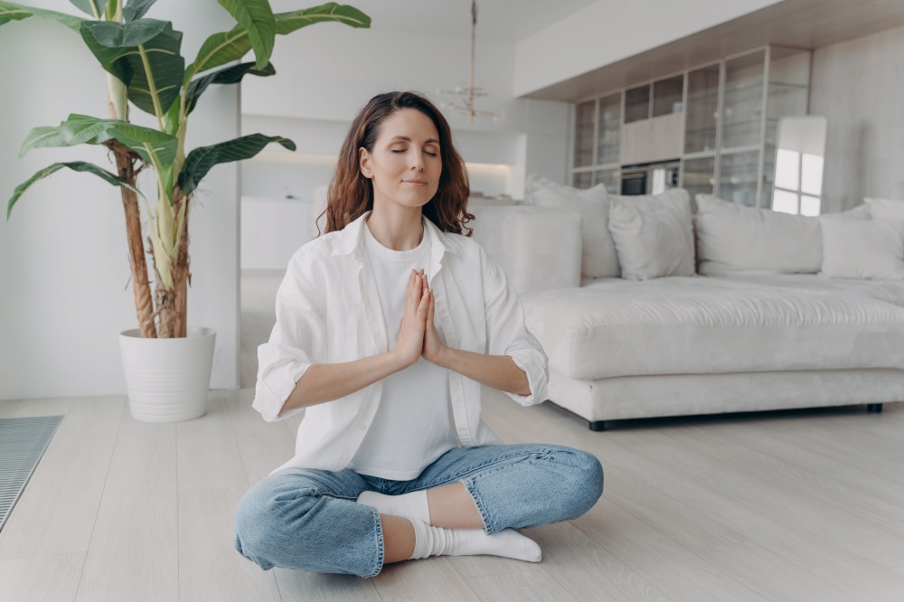Relaxed woman sit on floor alone in lotus position, folded hands in namaste gesture, practicing yoga, relieving stress. Serene hispanic female meditates breathing deep with closed eyes at home.. Relaxed woman meditates practicing yoga, relieving stress, breathing deep sitting on floor at home
