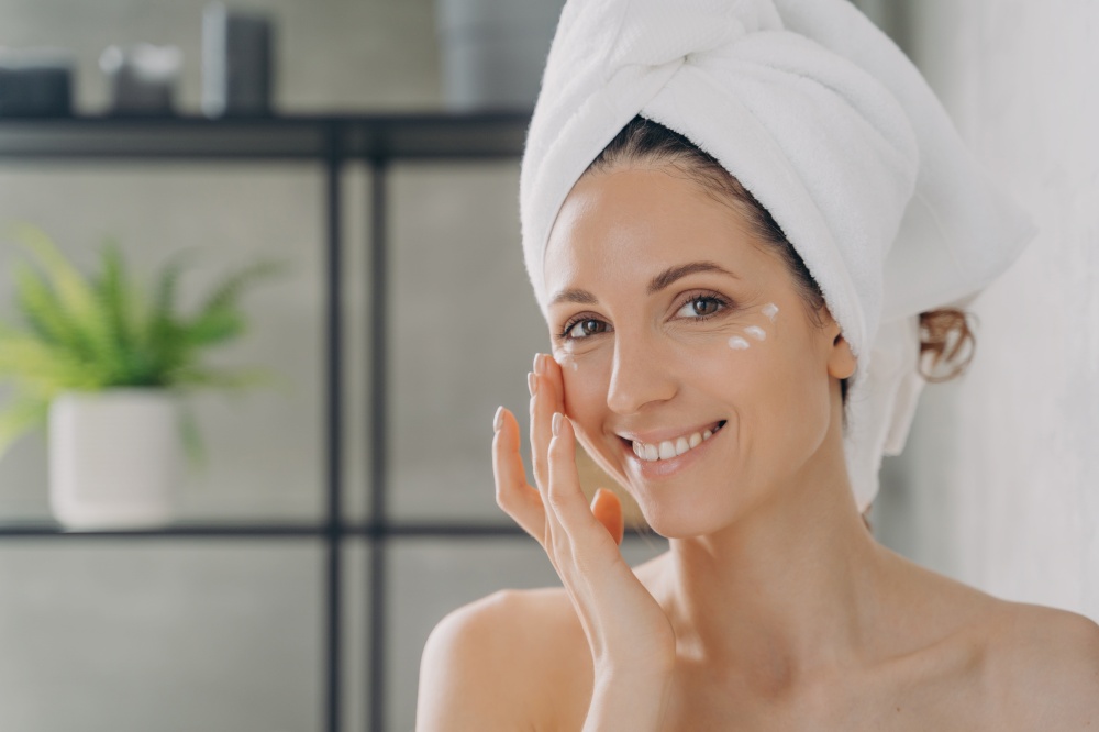 Smiling latina female with naked shoulders moisturizes under eye skin with white face cream, woman in towel apply moisturizing serum, looking at camera in bathroom, enjoying skincare routine at home.. Smiling latina female in towel moisturizes facial skin with cream in bathroom. Skincare treatment