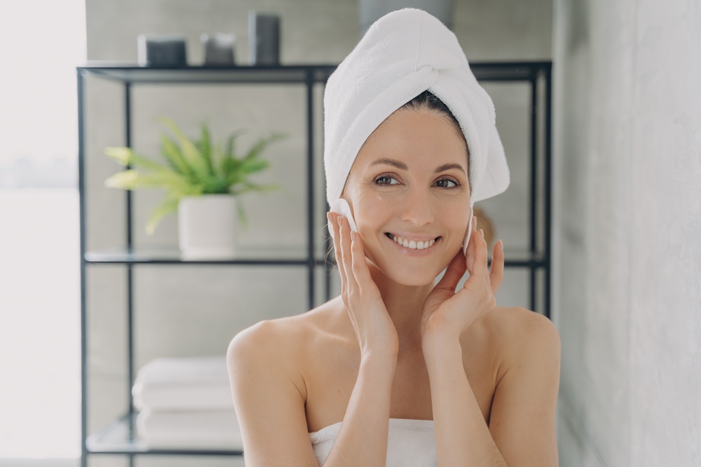 Female with towel on head cleanse face skin removing makeup using cotton pads after shower. Latina woman enjoying fresh clean healthy skin. Skincare beauty treatment, self care concept.. Happy female cleansing face skin removing makeup using cotton pads after shower. Skincare, self care