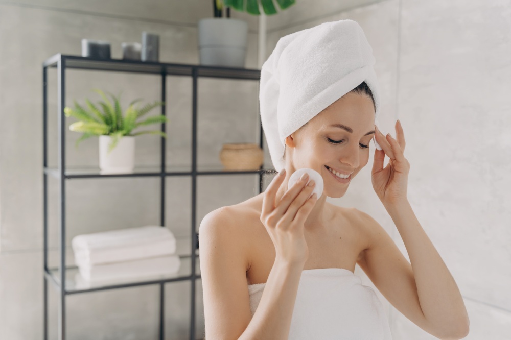 Smiling female cleanses facial skin by cotton pads in bathroom after shower. Woman wrapped in towel removing makeup by cleansing milk or micellar water, moisturizing face by tonic. Skincare routine.. Skin cleansing, skincare. Smiling female cleanse facial skin by cotton pads in bathroom after shower