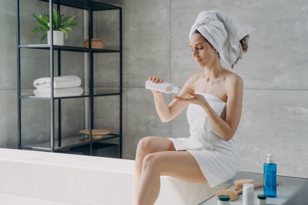 Young woman applying skincare anticellulite lotion doing body care morning routine in bathroom. Female holding bottle with natural cosmetics taking care of skin sitting on bath at home.. Female apply skincare anticellulite lotion doing body care routine in bathroom. Spa cosmetics ad