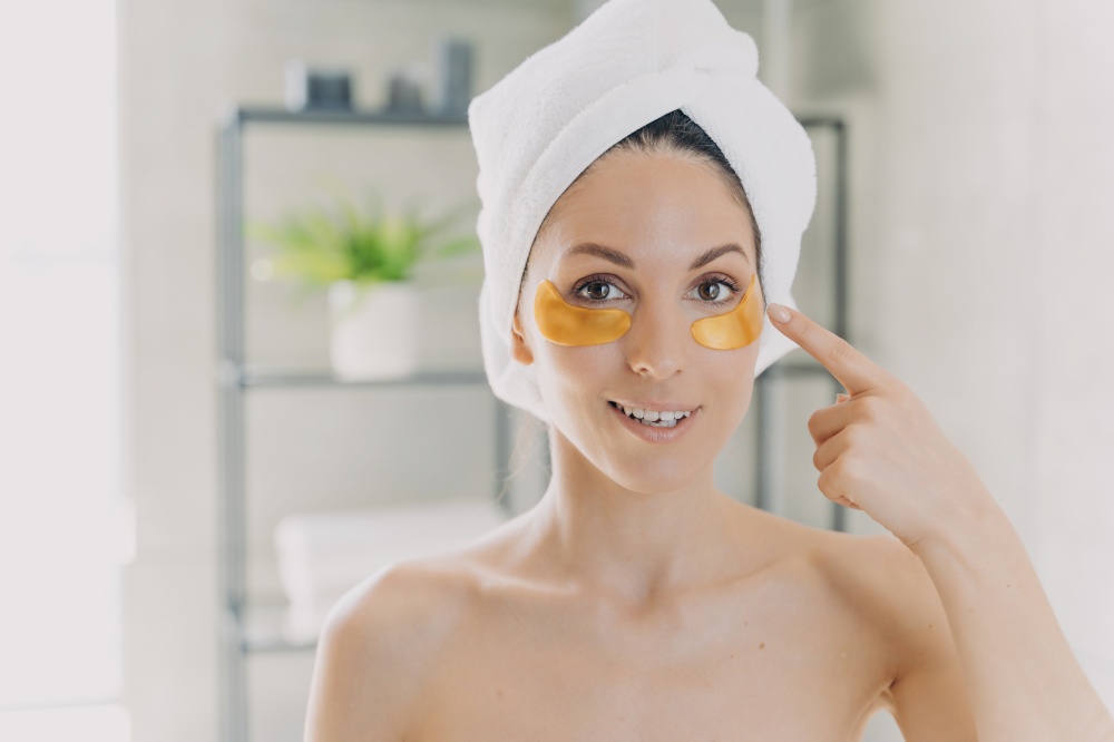 Hispanic woman with bare shoulders and towel on head shows under eye collagen golden pads, standing in bathroom. Skincare, antiaging moisturizing gold hydrogel patches, eye skin treatment.. Female shows under eye collagen golden patches standing in bathroom. Skincare, eye skin treatment