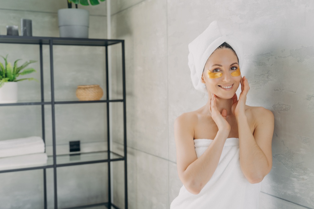 Smiling beautiful woman with moisturizing antiaging hydrogel patches under eyes, pretty female wearing white bath towel on head posing in bathroom during skincare treatment procedure.. Beautiful woman with antiaging hydrogel patches under eyes in bathroom. Skincare treatment procedure