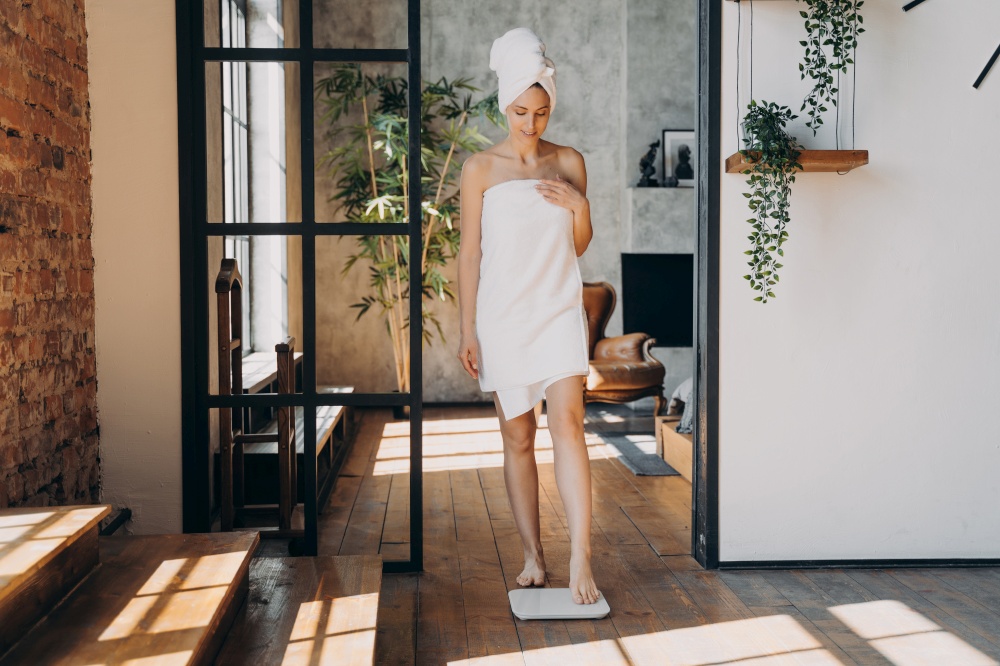 Female wrapped in towel after shower measures weight stepping onto electronic floor scales at home. Pretty young girl weighing in the morning. Slimming, wellness, body care concept.. Female wrapped in towel measure weight with scales at home in morning. Slimming, wellness, body care