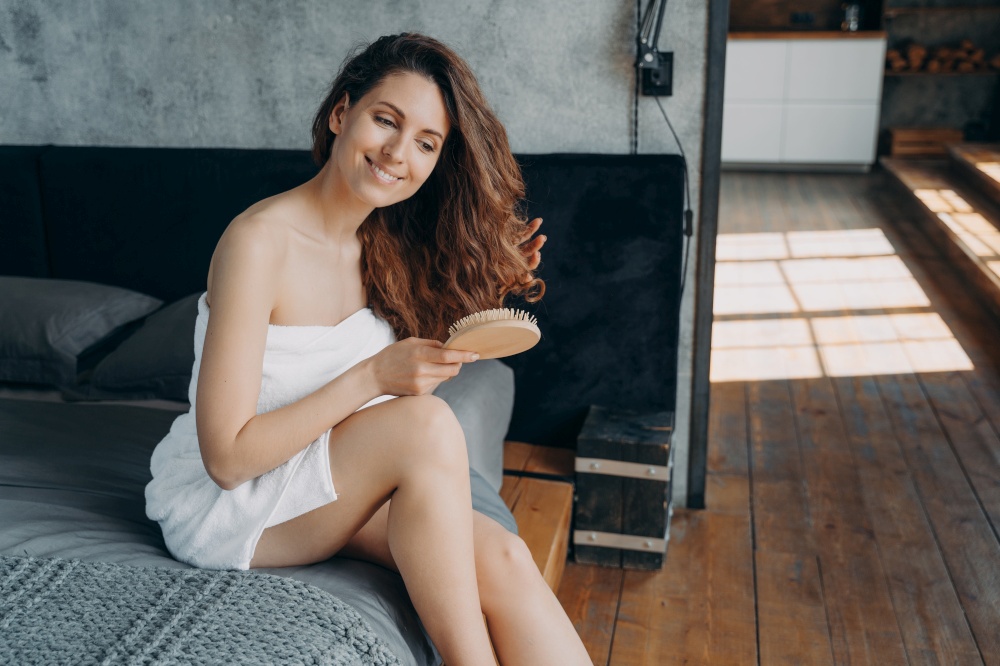 Smiling young spanish woman brushes long healthy hair with hairbrush after shower in bedroom. Girl wrapped in towel combing hairs, preening sitting on bed in the morning. Haircare, self-care routine.. Smiling girl brushes long healthy hair with hairbrush after shower in bedroom. Haircare cosmetics ad