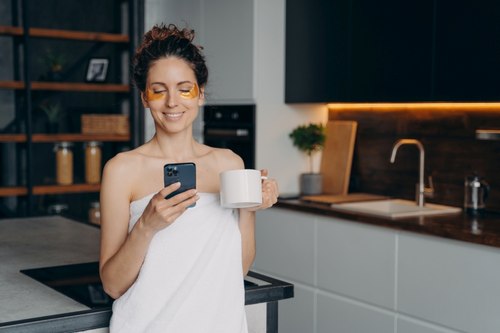 Modern young businesswoman with skincare under eye patches reads messages, news on smartphone, holding morning coffee cup. Happy girl wrapped in towel browsing social networks content after shower.. Businesswoman with skincare under eye patches reads message, news on smartphone after morning shower