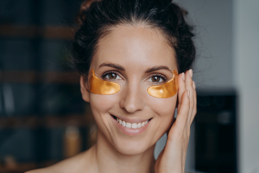 Smiling young latina woman applying moisturizing collagen golden under eye patches, happy pretty female does facial anti wrinkle beauty procedure at home, close up portrait. Skincare routine.. Smiling latina woman applying moisturizing golden under eye patches at home. Skincare beauty routine
