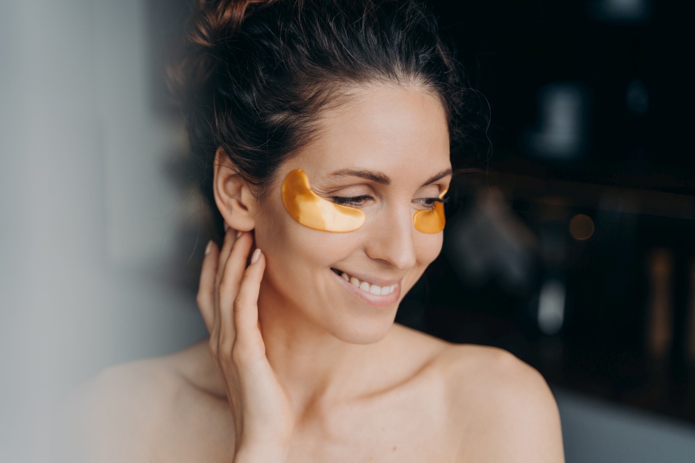 Smiling hispanic woman with bare shoulder applying hydrogel patches on under eyes skin, preventing wrinkles, moisturizing face. Happy girl enjoy skincare treatment procedure. Antiaging beauty routine. Pretty woman with hydrogel patches on under eyes skin. Skincare treatment, antiaging beauty routine