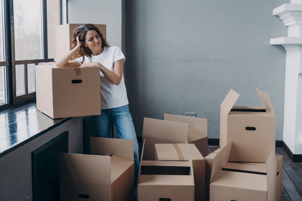 Spanish woman resting on moving day to new apartment, standing near window sill with boxes on floor. Female dreaming about happy future in own home. First realty, mortgage concept.. Female dreaming about new home on moving day standing with a lot of boxes. First realty, mortgage