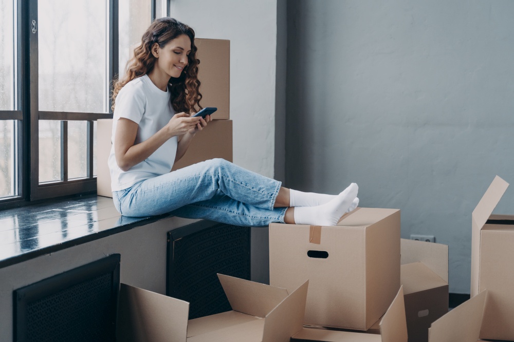 Modern girl homeowner tenant using smartphone app, choosing moving company, chatting with mover online, sitting on windowsill with cardboard boxes, waiting to move to a new house. Relocation concept.. Happy girl using smartphone apps, choosing moving company, sitting with boxes. Relocation, new home