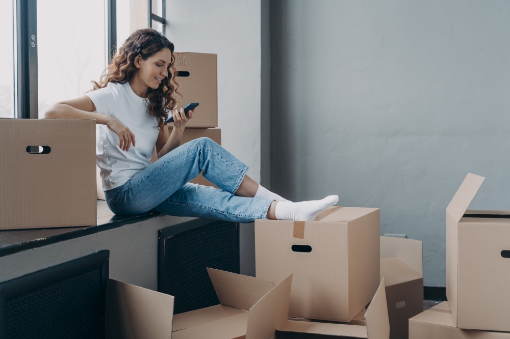 Smiling girl using smartphone apps, sitting on window sill with boxes on relocation day, happy female home buyer choosing moving service, or looking for interior design ideas, planing home renovation.. Girl holding phone sitting on windowsill with boxes selects moving service, planning home renovation