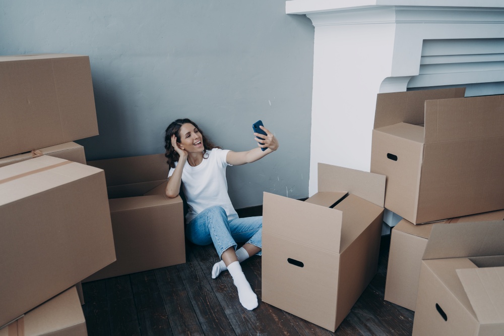 Happy female homeowner chatting by video call, holding smartphone, sitting with cardboard boxes. Girl communicate online during packing things for relocation, waiting for transportation service.. Joyful girl chatting by video call sitting with cardboard boxes during packing things for relocation