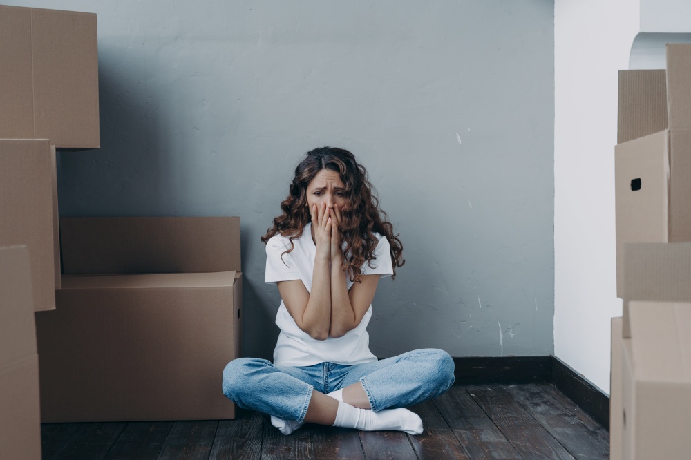Sad tired young hispanic girl feels stress due to moving, divorce or eviction sitting on the floor in empty room with cardboard boxes. Upset female tenant frightened of uncertain future and relocation. Sad tired girl feels stress due to moving, divorce or eviction sitting in room with cardboard boxes