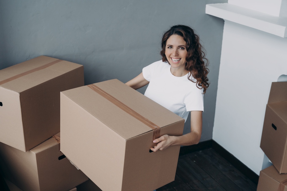 Joyful latina female homeowner holding cardboard box with personal things, moving in new house. Smiling woman carrying carton, packed belongings to home. Relocation and apartment renovation concept.. Joyful female homeowner standing in new house holding cardboard box. Moving, apartment renovation