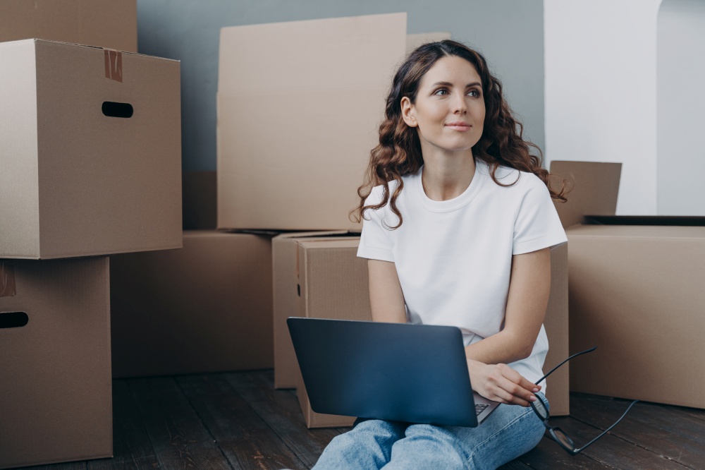Pensive spanish woman with laptop sit on floor near heap of cardboard boxes, search moving company on internet, buy furniture for home online, dreaming about new home on relocation day.. Happy girl with laptop sit on floor near cardboard boxes dreaming about new home. Relocation concept