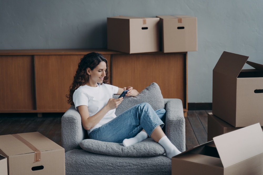 Moving day. Young spanish girl sitting in armchair between cardboard boxes, surfing information online on smartphone, shopping choosing furniture in internet store or ordering relocation service.. Moving day. Girl using phone app shopping furniture in online store sitting between cardboard boxes