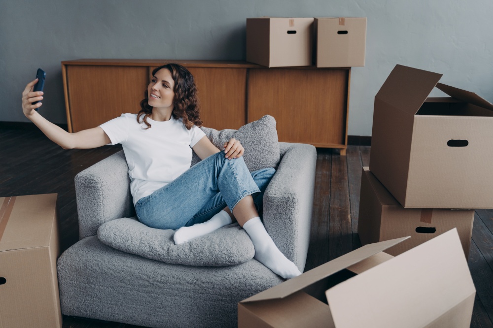 Smiling girl holding smartphone, chatting by video call in new apartment during relocation. Female communicates with friends or relatives, showing her house with cardboard boxes on moving day.. Girl holding phone communicates by video call, showing house with cardboard boxes on moving day