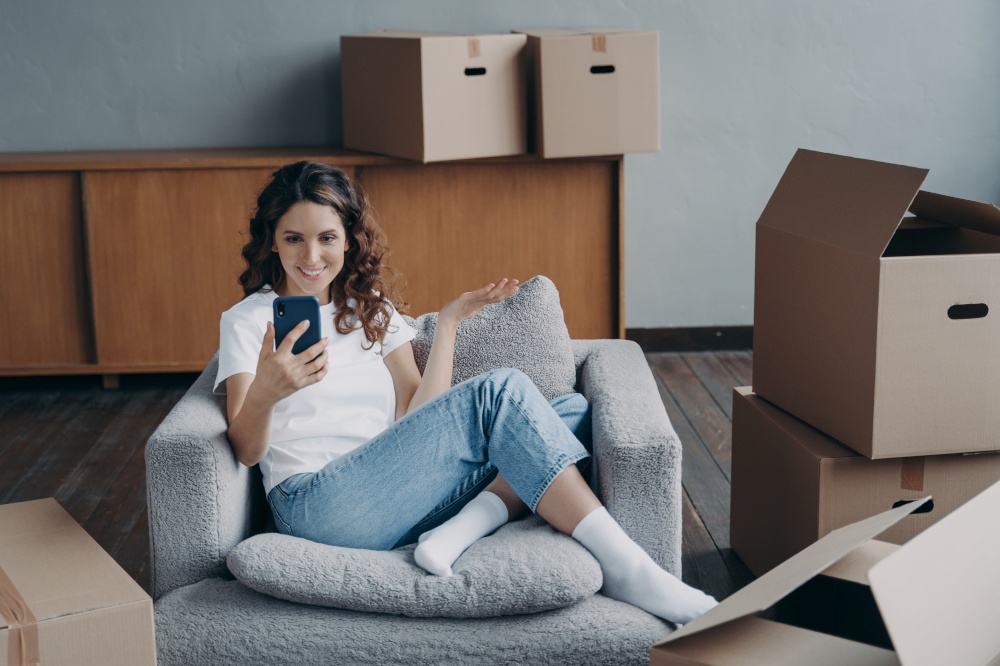 Happy young hispanic woman holding phone discussing new apartment design project, planing home renovation, chatting online by video call, sitting with boxes on moving day. Relocation concept.. Smiling girl discusses new house design, plans home renovation, chatting by video call on moving day