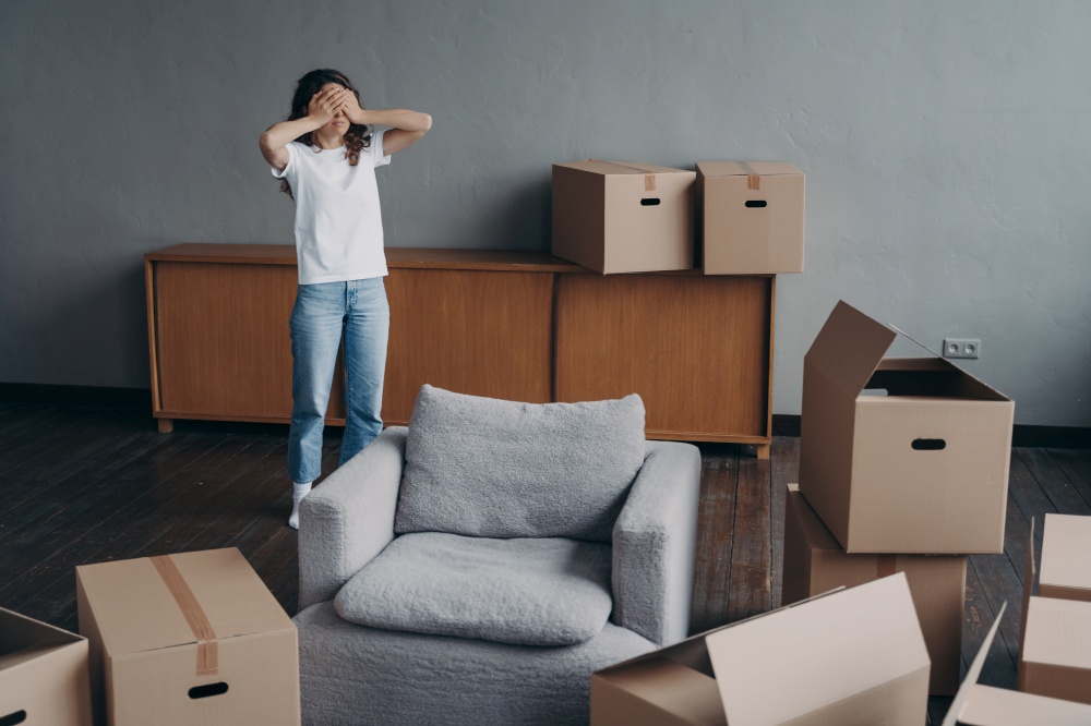 Tired woman surrounded by carton boxes having problem with packing things for removal, covers her eyes with hands. Exhausted girl feels stress of relocation day. Advertising of moving company.. Tired woman surrounded by carton boxes feels stress of relocation. Advertising of moving company