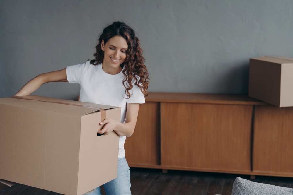 Happy spanish woman carrying cardboard box with belongings, relocating to first own apartment. Smiling female tenant brings carton with domestic things in living room on moving day. Mortgage, new home. Happy woman carrying cardboard box with things, relocating to first new house. Mortgage, relocation