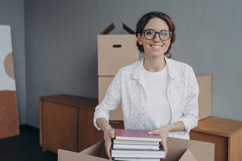 Smiling woman packing belongings in carton at home, happy hispanic female office worker wearing glasses puts things into cardboard box on moving day. New dwelling, relocation concept.. Smiling hispanic female office worker packing things on moving day. New dwelling, relocation concept