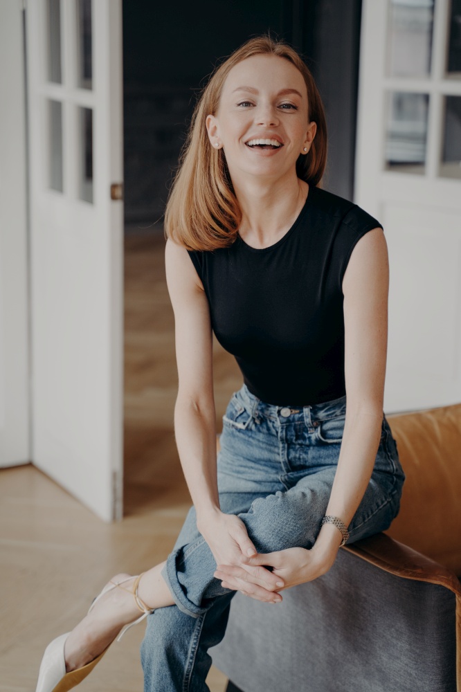 Lady is sitting on an armchair armrest and laughing. Emotional portrait of slim blonde tender woman. Redhead gorgeous lady in casual outfit is smiling. Happy european woman in apartment.. Lady is sitting on an armchair armrest and laughing. Emotional portrait of blonde tender woman.