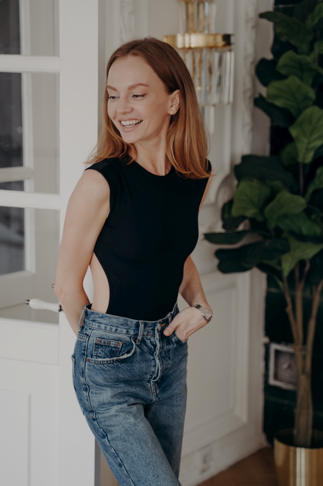 Attractive european lady in fashionable slim fit black bodysuit and jeans. Gorgeous sensual model is standing, looking aside and smiling. Luxurious apartment interior. Concept of beautiful body shape.. Attractive european lady in fashionable slim fit black bodysuit and jeans looking aside and smiling.