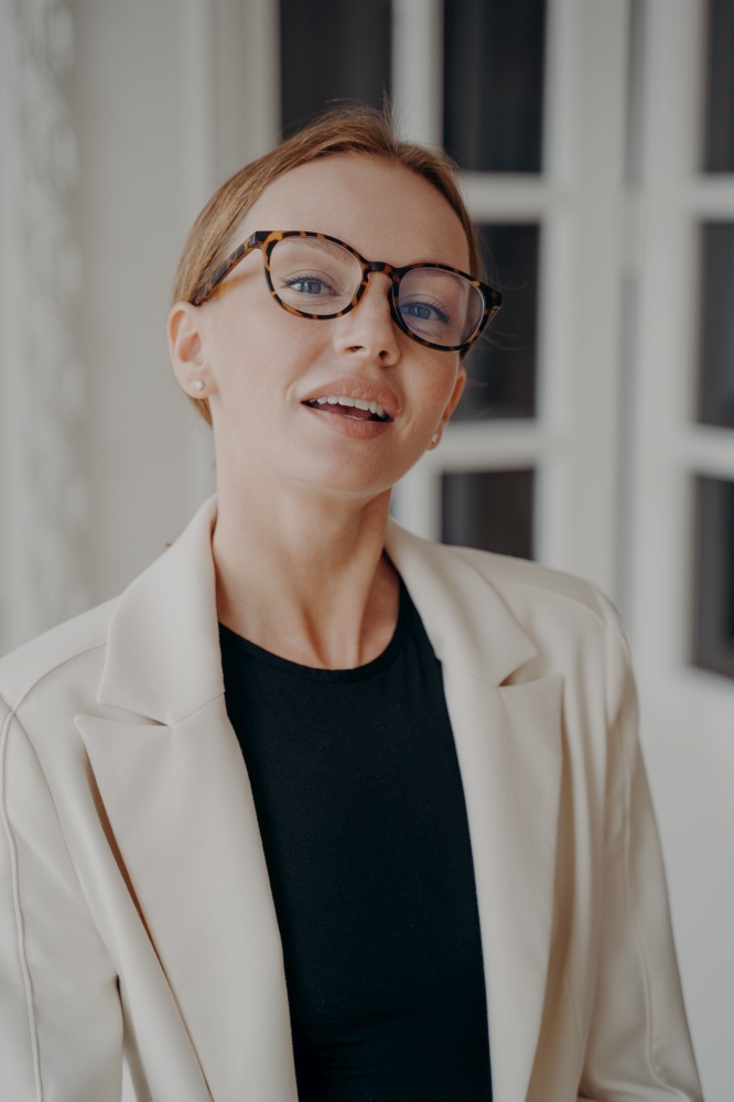 Portrait of stylish european businesswoman in glasses and white jacket posing in studio. Attractive blond lady in formal wear looks sensual. Expression of elegance, confidence and femininity.. Portrait of stylish businesswoman in glasses and white jacket. Lady in formal wear looks sensual.