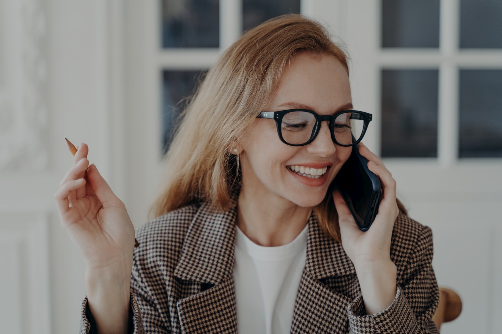 Elegant happy mid adult businesswoman in glasses is talking on phone. Pretty business lady has telephone call indoor. Attractive emotional european lady is freelancer or entrepreneur.. Elegant mid adult businesswoman in glasses is talking on phone. Lady is freelancer or entrepreneur.