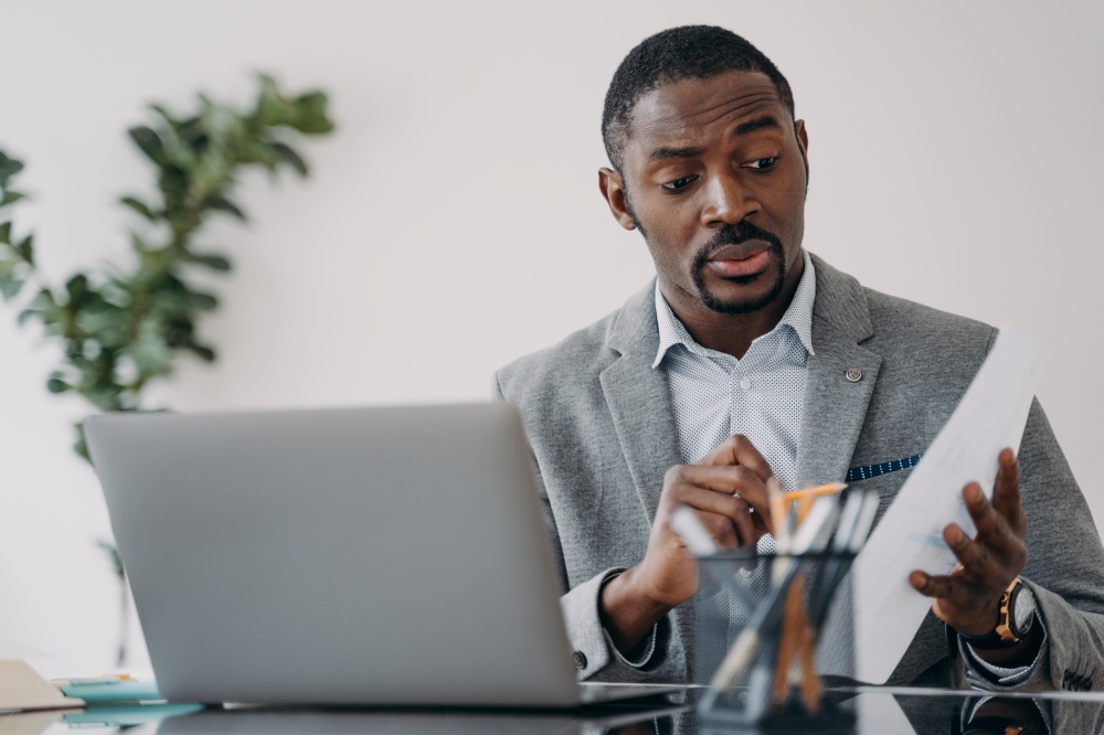 Puzzled frowning african american businessman discussing working project online by video call at laptop. Pensive dissatisfied black male manager solving work problem, holding paper report.. Puzzled african american businessman discussing working project online by video call at laptop