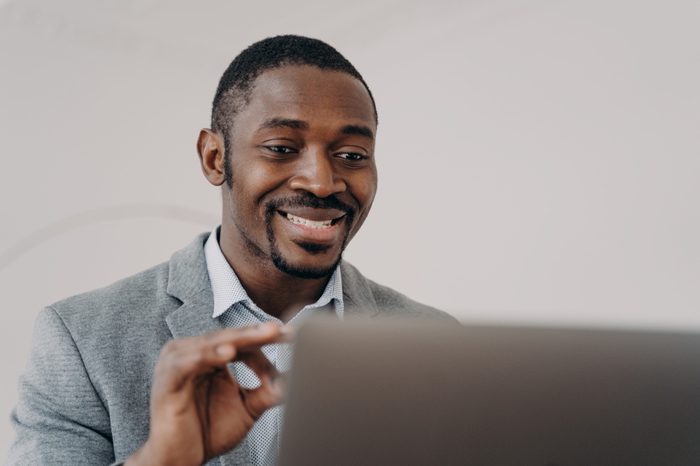 Smiling african american male manager talks by video call with client showing ok gesture. Friendly black businessman shows okay, looking at computer screen, consulting customer online at remote job.. Smiling african american male manager show ok gesture, talking with client by video call at laptop