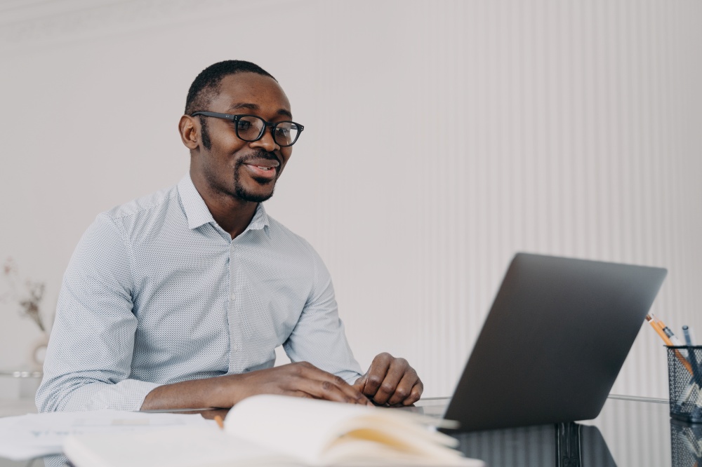 Smiling african american man wearing glasses working on laptop reading email with good news, sitting at office desk, satisfied with his business project. Black guy watching webinar, learning online.. Smiling african american man working on laptop on business project, reading email with good news